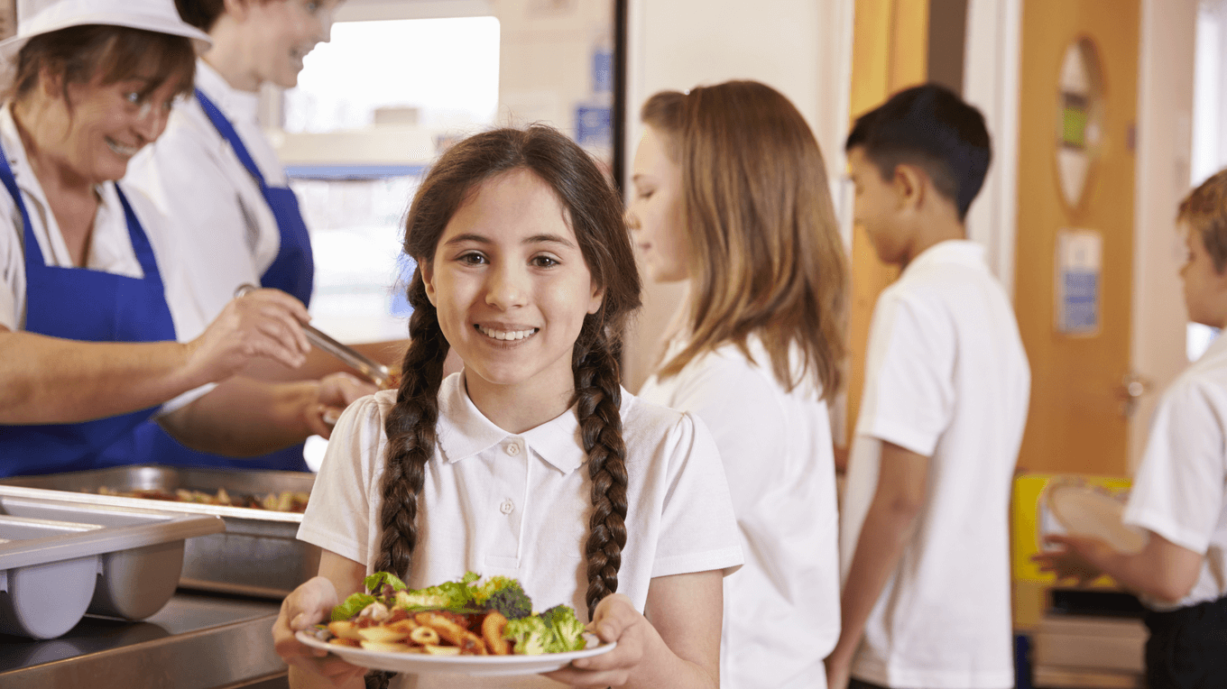 Шкільна їдальня - діти отримують порцію їжі, по центрі стоїть дівчина з тарілкою їжі у руках та посміхається.